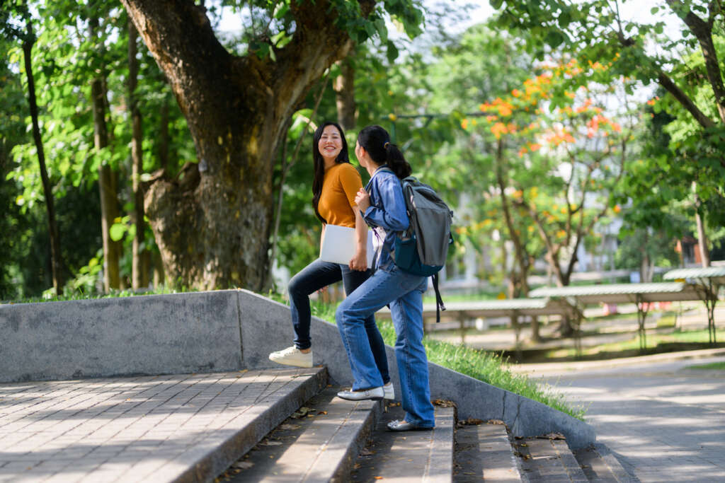 Two young adult female university students are walking up the stairs in a park, carrying laptop and backpack