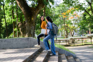 Two young adult female university students are walking up the stairs in a park, carrying laptop and backpack
